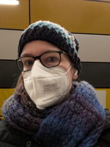 Eine Person mit Mütze, Brille und Maske. Am linken Ohr sieht man wie sie Airpods trägt.