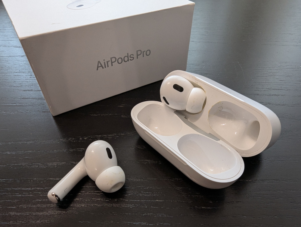 Zwei Airpod Kopfhörer mit Ladeschade und Verpackung. Einer der Kopfhörer steckt in der Ladeschale, einer liegt davor.