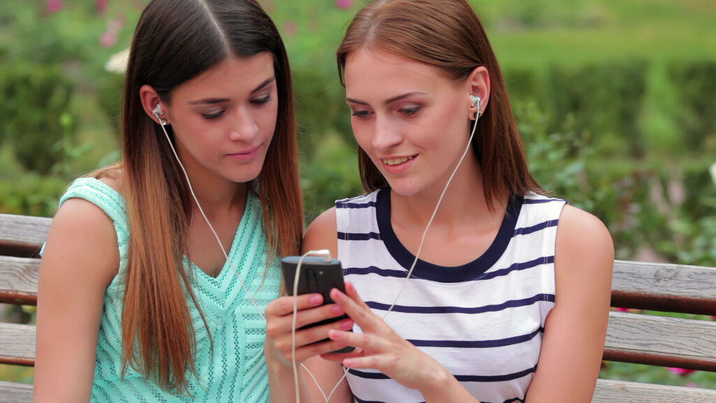 Zwei junge Frauen, die von einem Handy zusammen mit Ohrenstöpselkopfhörern etwas hören