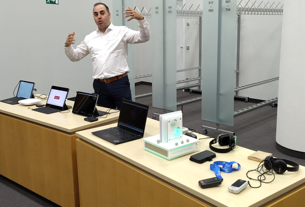 Yaser Georgos von Resound hinter zwei Tischen mit verschiedenen Laptops und anderen Geräten die Auracast senden oder empfangen